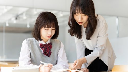 机で教材を見ながら学ぶ生徒と、隣で説明する大人